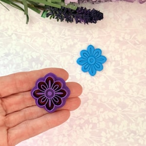 May include: A hand holds a purple flower-shaped cookie cutter with a detailed floral design. A separate blue flower-shaped cutter is also visible. The cutters are likely for baking or crafting, with a floral theme.
