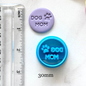 May include: A purple polymer clay charm with the text "DOG MOM" and a paw print. A blue plastic cookie cutter with the same text and paw print. The cookie cutter is 30mm in diameter.