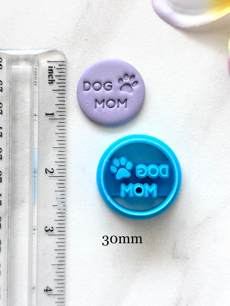 May include: A purple polymer clay charm with the text "DOG MOM" and a paw print. A blue plastic cookie cutter with the same text and paw print. The cookie cutter is 30mm in diameter.