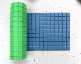 Può includere: Un mattarello in silicone verde e blu con un motivo a griglia quadrata. Il mattarello è steso su una superficie bianca.
