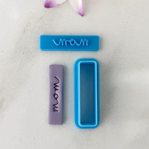 May include: A set of two blue plastic cookie cutters. One cutter is shaped like a rectangle and has the word "mom" embossed on it. The other cutter is a rectangle with a smaller rectangle cut out of the center.