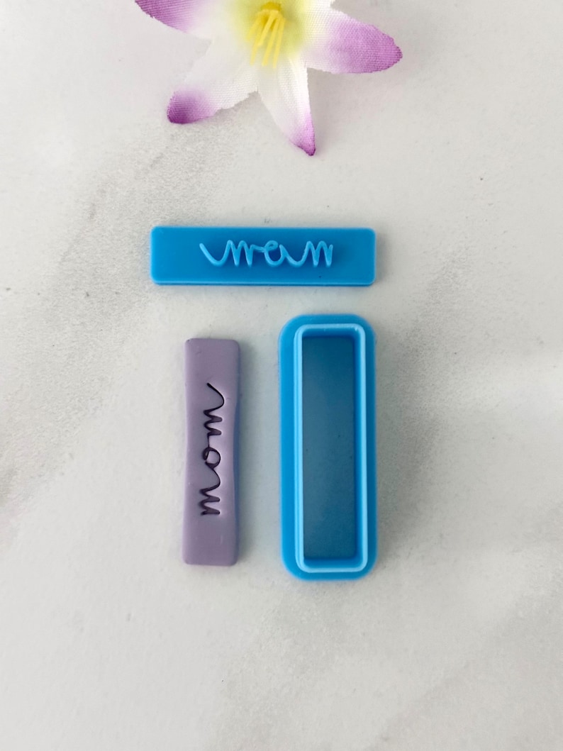 May include: A set of two blue plastic cookie cutters. One cutter is shaped like a rectangle and has the word "mom" embossed on it. The other cutter is a rectangle with a smaller rectangle cut out of the center.