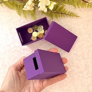 May include: A purple box with a matching lid and a small rectangular opening. Inside the open box are several daisy-shaped charms in various colors. The box is held in a hand.