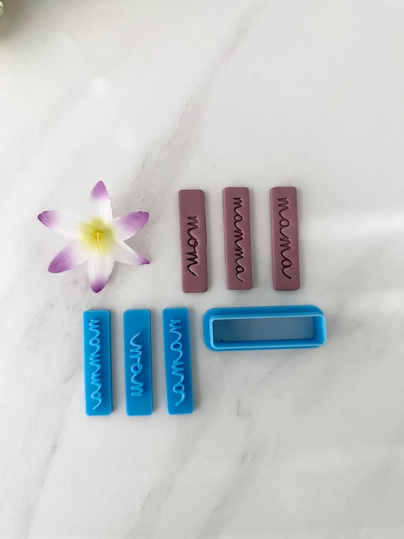 May include: A set of nine blue and purple polymer clay stamps with the words "mom", "mamma", "mama", "mommy", "momma", and "mommy" in a cursive font. The stamps are arranged on a white marble surface.