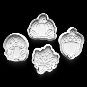 May include: Four white plastic cookie cutters in autumn shapes: a pumpkin, a mushroom, a maple leaf, and an acorn. The cutters are arranged against a black background, showcasing their detailed designs.