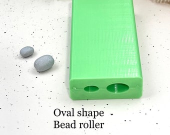 May include: Green plastic bead roller with two oval-shaped molds. The roller is on a white surface with two gray oval-shaped beads.