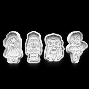 May include: Four white plastic cookie cutters in the shapes of characters from The Nutcracker ballet. The shapes include a girl, a nutcracker soldier, a mouse king, and a ballerina. The cutters are on a black background.