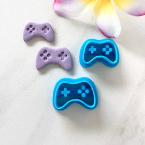 Może przedstawiać: Dwa fioletowe plastikowe wykrawacze do ciasteczek w kształcie kontrolerów do gier wideo i jeden niebieski plastikowy wykrawacz do ciasteczek w kształcie kontrolera do gier wideo.