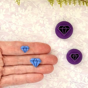May include: A light blue diamond-shaped charm and two purple circular charms with a black diamond design. The charms are displayed on a floral patterned background.