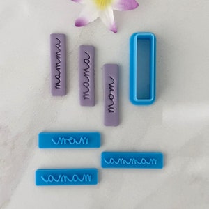 May include: A set of six blue and purple polymer clay cookie cutters with the word "mama" written in different styles. The cutters are on a white surface with a white flower.