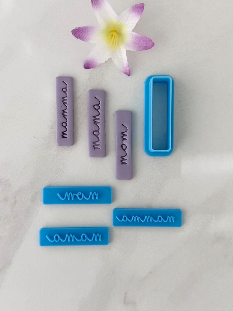 May include: A set of six blue and purple polymer clay cookie cutters with the word "mama" written in different styles. The cutters are on a white surface with a white flower.