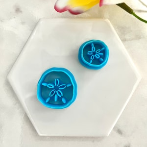 Könnte beinhalten: Zwei blaue Plastik-Ausstecher in Form einer Sanddollar mit einem Sternburst-Design in der Mitte. Die Ausstecher befinden sich auf einem weißen sechseckigen Teller.