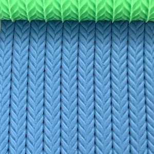 May include: A close-up of two textured silicone rolling pins. The top rolling pin is green and the bottom rolling pin is blue. Both rolling pins have a herringbone pattern.