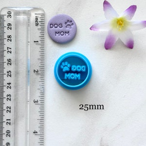 May include: A blue plastic cookie cutter with the text "DOG MOM" and a paw print. The cutter is 25mm in diameter. A purple fondant cookie with the same text and paw print is shown next to the cutter.
