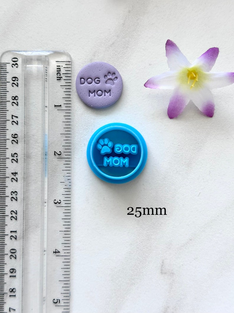 May include: A blue plastic cookie cutter with the text "DOG MOM" and a paw print. The cutter is 25mm in diameter. A purple fondant cookie with the same text and paw print is shown next to the cutter.