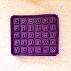 May include: A purple alphabet and number stamp set. The set includes lowercase letters, numbers, and symbols arranged in a square tray. The letters and numbers are raised and ready for use. The tray is a solid, vibrant purple color.