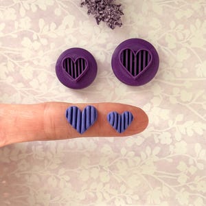 May include: Purple heart-shaped cookie cutters and matching heart-shaped cookies. The cutters are circular with a heart-shaped cutout and vertical lines. The cookies are held on a finger. A sprig of dried lavender is in the background.