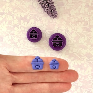 May include: Two purple cupcake-shaped stamps and two blue cupcake-shaped objects. The stamps have a cupcake design with a heart in the center. The blue objects are held in a hand. A sprig of purple flowers is in the background.
