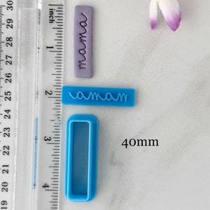 May include: Two blue plastic cookie cutters, one with the word "mama" and the other a rectangular shape. The cutters are on a white surface with a ruler in inches and a purple polymer clay stamp with the word "mama" on it.