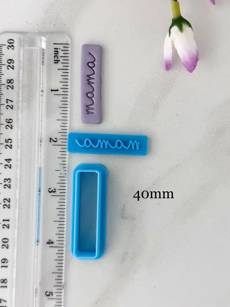 May include: Two blue plastic cookie cutters, one with the word "mama" and the other a rectangular shape. The cutters are on a white surface with a ruler in inches and a purple polymer clay stamp with the word "mama" on it.