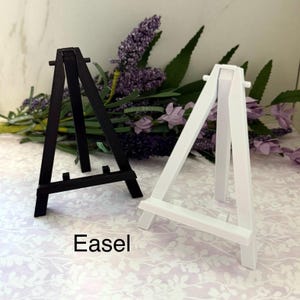 Puede incluir: Se muestran dos pequeños caballetes de madera, uno negro y otro blanco. La palabra "Easel" está impresa debajo. Los caballetes tienen forma triangular y tienen una pequeña repisa para sostener un lienzo o una obra de arte.