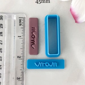 May include: A set of three blue and purple plastic cookie cutters. The cutters are shaped like rectangles and feature the word "mom" in a cursive font. The cutters are 45 mm in size.