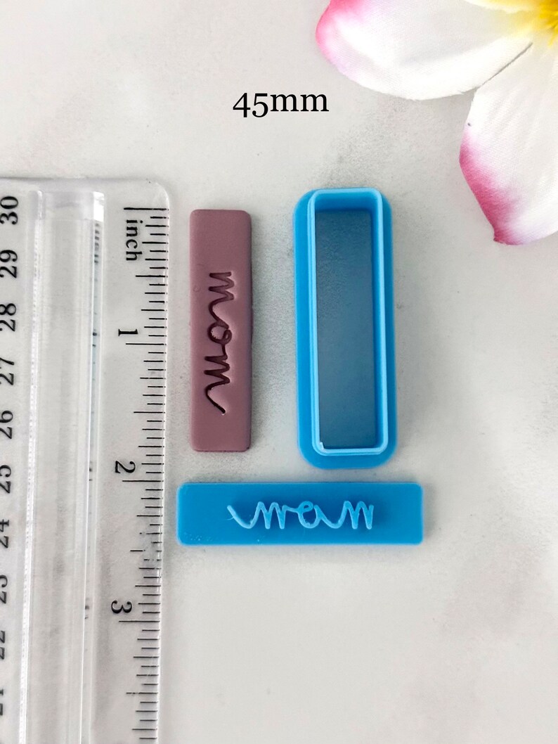 May include: A set of three blue and purple plastic cookie cutters. The cutters are shaped like rectangles and feature the word "mom" in a cursive font. The cutters are 45 mm in size.