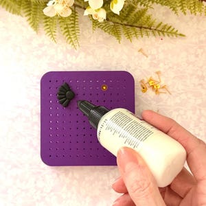 May include: A purple pegboard with a black decorative element and a bottle of glue. The pegboard has small holes and a gold-coloured stud. A hand holds the glue bottle, ready for crafting.