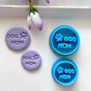 May include: Two purple clay cookie cutters with the text "Dog Mom" and a paw print. Two blue plastic cookie cutters with the text "Dog Mom" and a paw print.