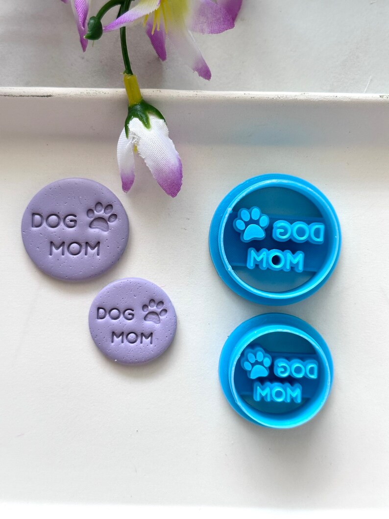 May include: Two purple clay cookie cutters with the text "Dog Mom" and a paw print. Two blue plastic cookie cutters with the text "Dog Mom" and a paw print.