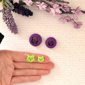 May include: Two purple cat-shaped cookie cutters and two green cat-shaped decorations. The cookie cutters have a cat face design inside a circular frame. The green decorations are held in a hand and have a textured appearance.