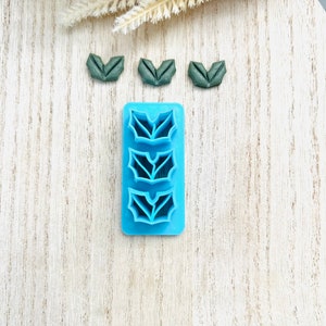 Könnte beinhalten: Eine blaue Plastikform mit drei Formen von Stechpalmenblättern. Drei grüne Stechpalmenblätter liegen auf einer weißen Holzoberfläche.