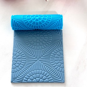 May include: A blue silicone rolling pin with a geometric pattern. The rolling pin is used to create decorative patterns on fondant or clay.