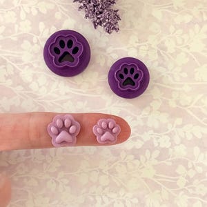 Puede incluir: Cortadores de galletas morados con huellas de patas y adornos con forma de huellas de patas. Los cortadores de galletas son circulares y tienen un diseño de huella de pata en relieve. Los adornos son de color rosa claro y tienen forma de huella de pata.