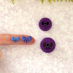 May include: A selection of purple and blue bow-shaped craft tools. Two small bows are held by a finger, while two larger bow-shaped stamps are on a floral patterned surface. The stamps are circular with a bow design.