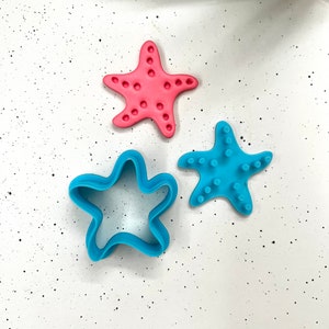 May include: Three star-shaped cookie cutters, two blue and one pink. The blue cutters are plain, while the pink cutter has a textured surface.