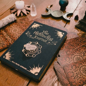 May include: A navy blue journal with gold lettering that reads "The Pathfinding Journal" and a gold moon and star design. The journal is on a wooden table with a deck of tarot cards, a white candle, and other crystals.