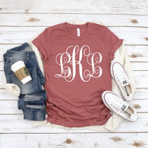May include: A maroon t-shirt with a white monogram of the letters "BKL" in a cursive font. The shirt is laid flat on a white wooden surface with a pair of white canvas shoes and a pair of blue jeans.