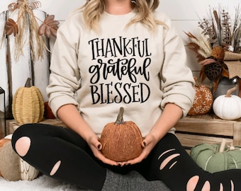 Peut inclure: Une femme portant un sweat-shirt beige avec le texte "Thankful grateful blessed" en lettres noires. Elle tient une petite citrouille orange scintillante dans ses mains. L'arrière-plan présente une décoration d'automne, y compris des citrouilles, des courges et une lanterne.
