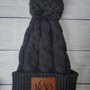 Op de afbeelding: Grijze kabelgebreide beanie met een pluche pompon en een bruin leren patch met het monogram "M&S" in zwart.