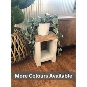 May include: A small wooden plant stand with a white painted base and a natural wood top. A white ceramic pot with a trailing green plant sits on top. The text "More Colours Available" is at the bottom.