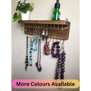 May include: Wooden jewellery organiser with gold hooks. The organiser has a shelf on top and is mounted on a wall. The shelf has a small green plant on it. The organiser is holding necklaces, earrings and bracelets.