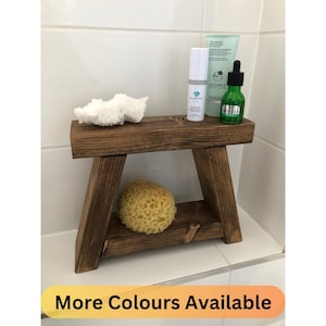 May include: A small, dark brown wooden stool with a rectangular top and angled legs. On the top surface sits a white cylindrical bottle, a light green tube labelled Liz Earle, and a green glass dropper bottle. A natural yellow sponge rests below.