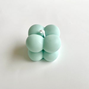 May include: A mint green bubble candle with a square shape. The candle is made of several spherical shapes joined together, with a white wick centered on top. The candle is a decorative item.