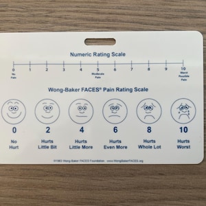 May include: A white card with a hole at the top, featuring the "Numeric Rating Scale" and the "Wong-Baker FACES Pain Rating Scale." The scale ranges from 0 to 10, with corresponding facial expressions and pain descriptions. The card measures approximately 5 inches by 3 inches.