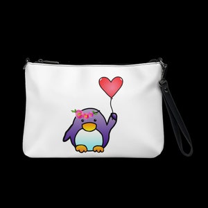 May include: White clutch with a black zipper and wrist strap. The clutch features a cartoon penguin with a floral crown holding a red heart-shaped balloon. The penguin is purple, with a white belly and yellow feet.