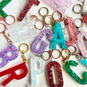 May include: A collection of colorful glitter resin letter keychains. The letters are in various colors, including pink, blue, green, red, and white. Each letter is attached to a silver key ring.