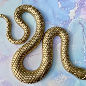 May include: A gold-colored snake sculpture with a textured, scaly surface. The snake is curled in a figure-eight shape.