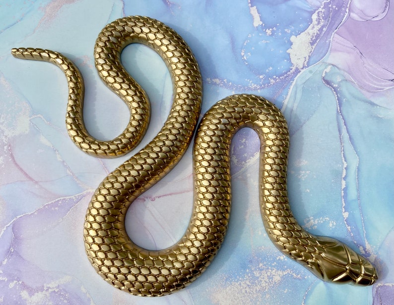 May include: A gold-colored snake sculpture with a textured, scaly surface. The snake is curled in a figure-eight shape.