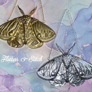 May include: Two metal moth ornaments, one gold and one silver, hang from chains. The moths have detailed wing patterns and are displayed against a marbled background. The text "Flutters & Stitch" is written in cursive.